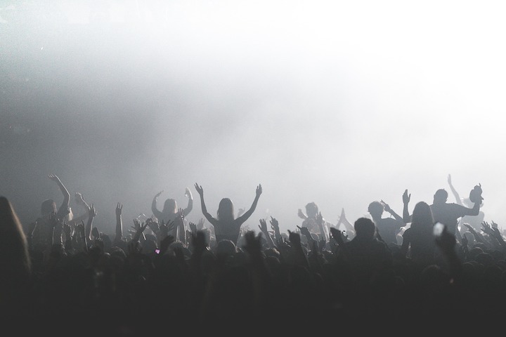 Backs of audience in the foreground and smoke from a machine lit by stage lights.